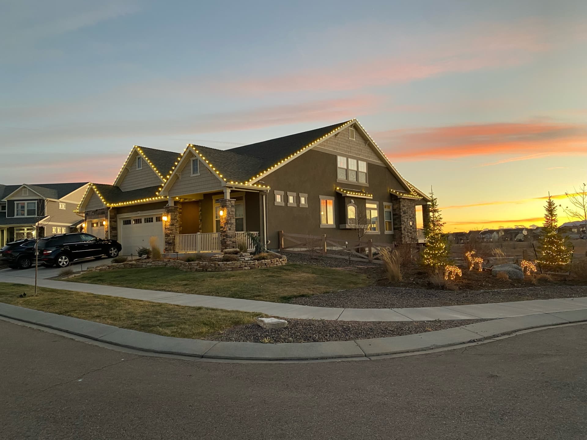 Craftsman home with warm white lights along every roofline at sunset, mini-lit trees and glowing reindeer in the side yard